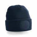 Product BC446 by Beechfield is a dark blue knit beanie featuring a wide cuff and a textured circular patch on the front. The ribbed pattern of the fabric lends it a cozy and snug look, set against a white background for display.