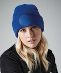 A person with long blonde hair is wearing a bright blue Beechfield BC446 and a black denim jacket. The beanie has a round patch on the front. They have a neutral expression and are set against a gray background.