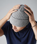 A person wearing a navy blue polo shirt and a gray Beechfield BC447 knit beanie cap holds the top of the beanie with both hands. The person's head is slightly bowed, and they are wearing a bracelet on their left wrist. The background is plain and light-colored.