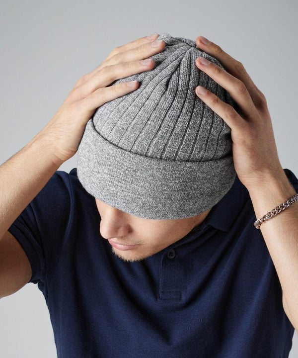 A person wearing a navy blue polo shirt and a gray Beechfield BC447 knit beanie cap holds the top of the beanie with both hands. The person's head is slightly bowed, and they are wearing a bracelet on their left wrist. The background is plain and light-colored.