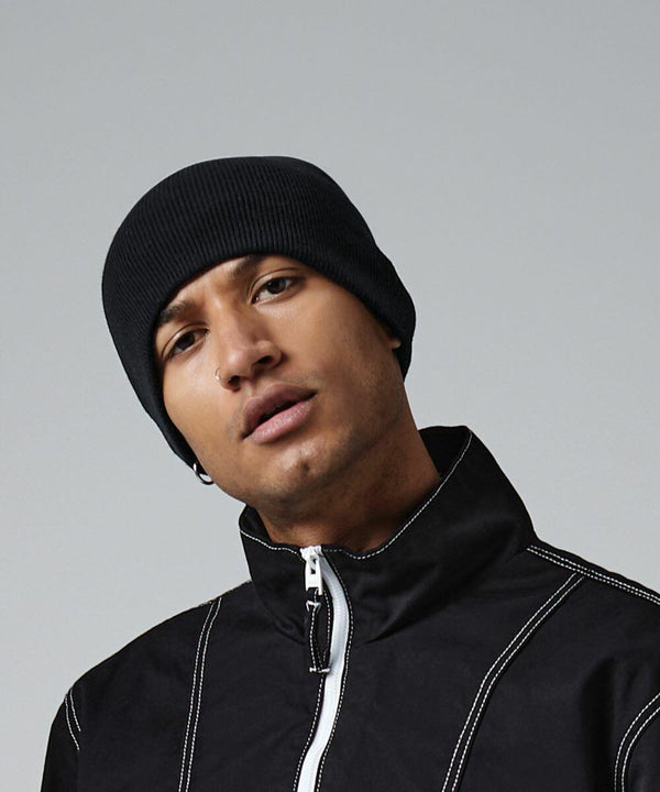 A person stands wearing a black Beechfield BC44R beanie and a black zip-up jacket with white stitching. They have stud earrings and a neutral expression, facing the camera with a light gray background.