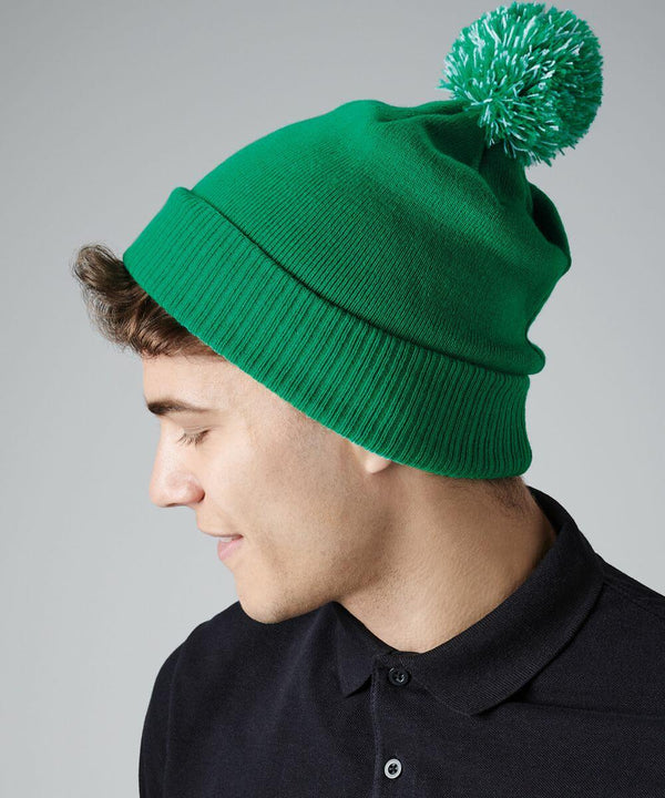 Young man in profile wearing a bright green Beechfield BC450 knit beanie with a large green and white pom-pom on top. He has short brown hair and is dressed in a black collared shirt. The background is a plain grey.