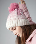 A person with long brown hair wears a Beechfield BC459, a pink and white knit beanie with a pink pom-pom. They are looking down and wearing a light pink jacket. The background is plain and gray, giving a neutral setting.