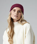 A person with long, wavy blonde hair wears a burgundy Beechfield BC45N beanie and a white turtleneck sweater. The background is a plain light gray color. The person gazes slightly off to the side with a neutral expression.