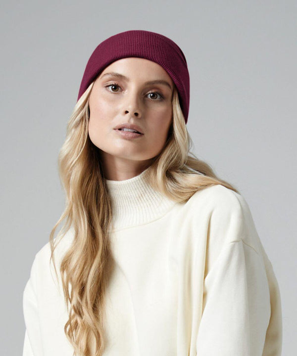 A person with long, wavy blonde hair wears a burgundy Beechfield BC45N beanie and a white turtleneck sweater. The background is a plain light gray color. The person gazes slightly off to the side with a neutral expression.