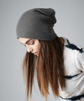 A person with long, straight brown hair is wearing a gray Beechfield BC461 beanie and a white tie-dye shirt with black accents. They are looking downward against a plain gray background.