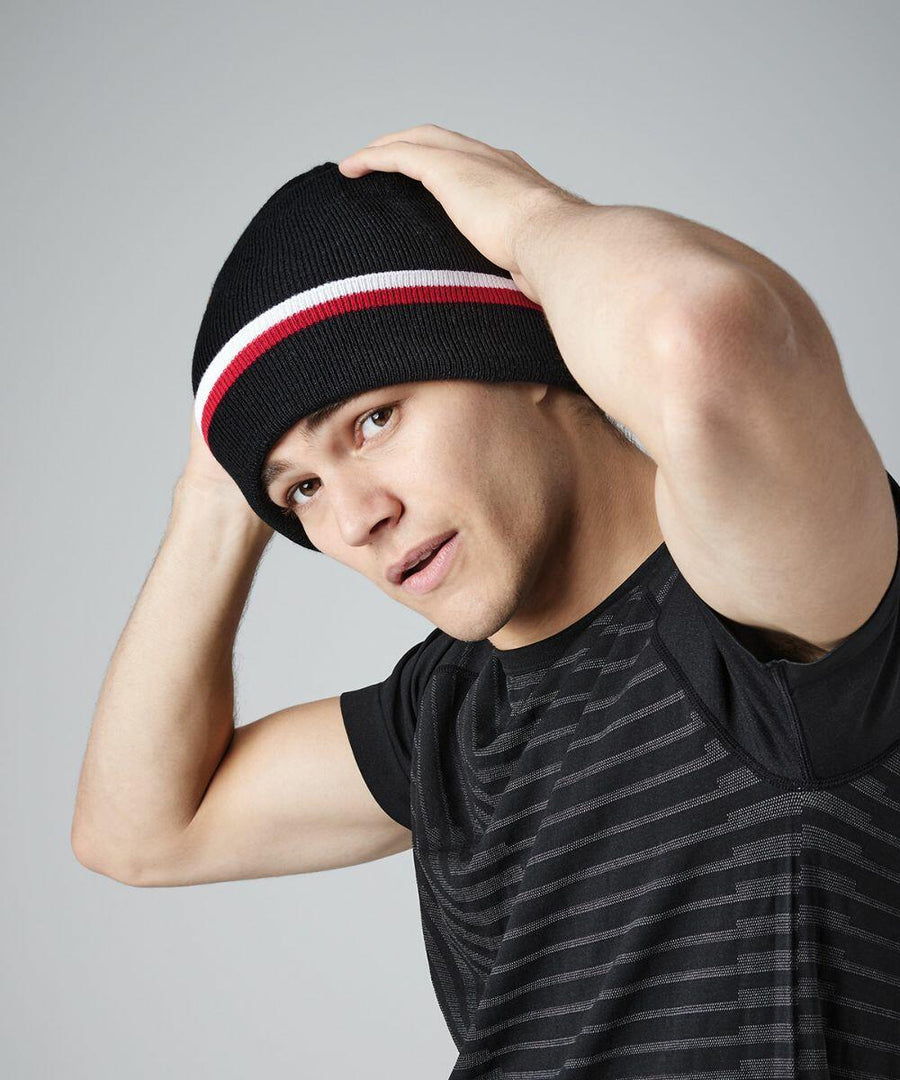 A person wearing a black t-shirt and a Beechfield BC471 beanie with red and white stripes adjusts their hat with both hands. They have a neutral expression and are looking slightly to the side. The background is plain gray.