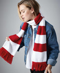 A person wearing a white and red striped Beechfield BC479 scarf with fringed ends, a denim jacket, and a white shirt. The individual's head is slightly tilted down, with short, light hair parted to one side. They have a calm expression against a gray background.