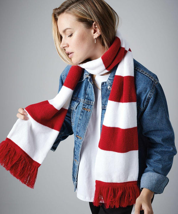 A person wearing a white and red striped Beechfield BC479 scarf with fringed ends, a denim jacket, and a white shirt. The individual's head is slightly tilted down, with short, light hair parted to one side. They have a calm expression against a gray background.