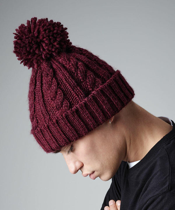 A person with closely-cropped hair, wearing a maroon Beechfield BC480 knitted hat with a pom-pom on top, looks downward. They are dressed in a black garment and positioned against a plain grey background.