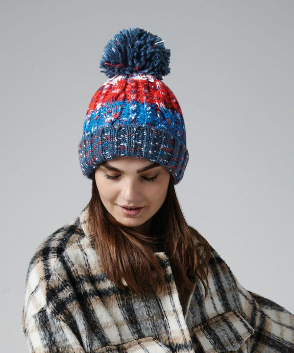 A person with long hair is wearing a colorful knit beanie from Beechfield, specifically the BC486, with a pom-pom on top. The beanie features red, blue, white, and gray colors. They are also wearing a black and white checkered coat and looking downward slightly. The background is plain gray.