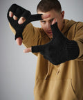 A person wearing a beige hoodie and black Beechfield BC491 fingerless gloves is posing with their hands extended forward, fingers forming a frame. They are looking through the makeshift "frame" created by their hands. The background is plain gray.