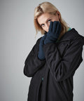 A person with blonde hair wears a black BC495 Beechfield jacket and holds their face in both hands, covered with dark gloves. They gaze directly at the camera with a neutral expression against a plain grey background.