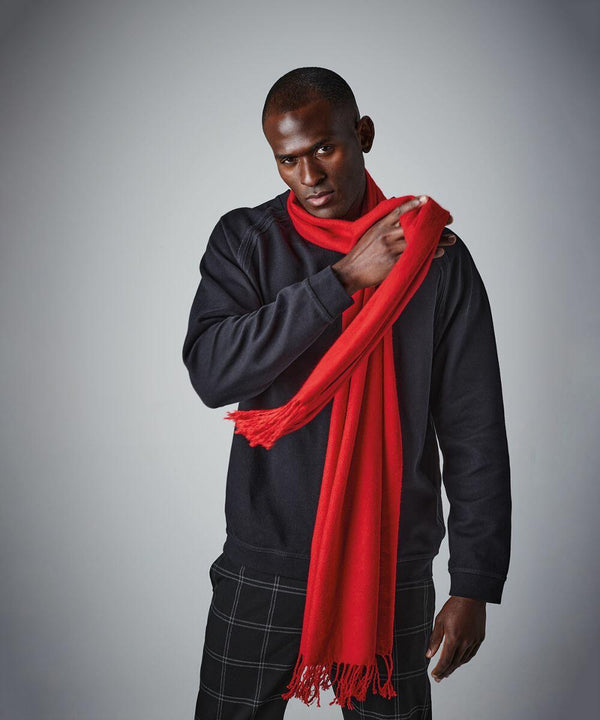 A man is posing against a gray background while adjusting a bright red Beechfield BC500 draped over his shoulder. He is wearing a black sweatshirt and patterned dark pants. He looks directly at the camera, holding one end of the Beechfield BC500 in his hand.