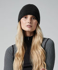 A person with long, wavy blond hair wears a black Beechfield BC501 beanie and a black zip-up top with white striped sleeves. The background is a plain, light gray color. The individual gazes directly at the camera with a neutral expression.
