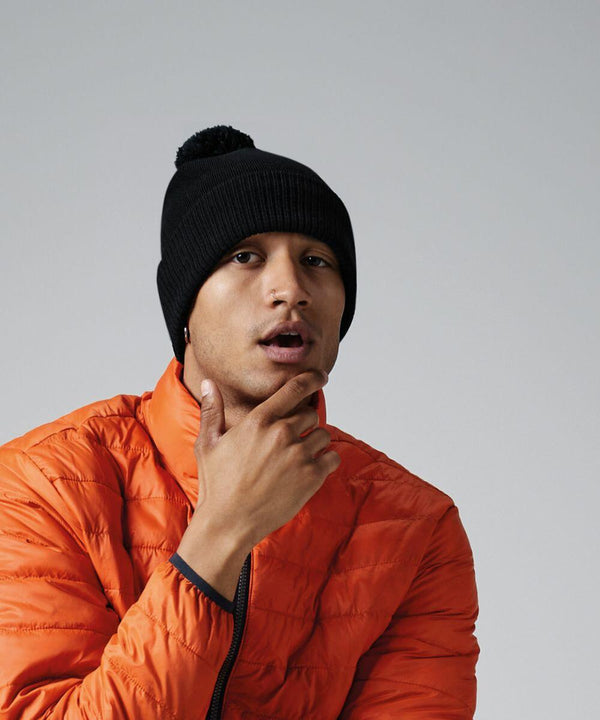 A person wearing a black Beechfield BC502 beanie and an orange puffer jacket with a blue zipper is posing with their right hand touching their chin and their mouth slightly open against a plain gray background.