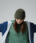 A person with long brown hair is looking down while wearing a green knit beanie (Beechfield BC505), a green hoodie, and a blue and white jacket. The individual’s arms are slightly outstretched, and the background is plain grey.
