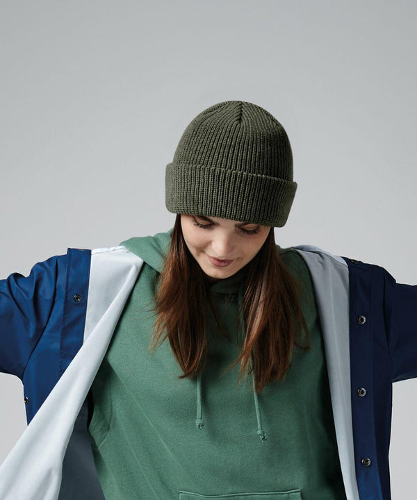 A person with long brown hair is looking down while wearing a green knit beanie (Beechfield BC505), a green hoodie, and a blue and white jacket. The individual’s arms are slightly outstretched, and the background is plain grey.
