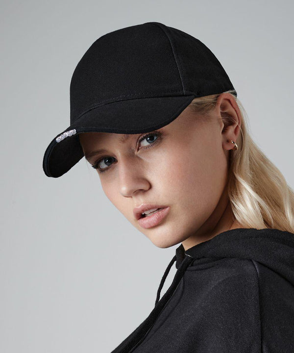 A person with long blonde hair is wearing a black BC515 baseball cap by Beechfield and a black hoodie. They have a neutral expression, and the background is plain gray. A small part of an earring is visible on their ear.