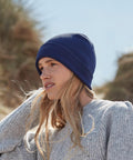 A person with long blonde hair wearing a blue Beechfield BC51N and a gray knit sweater looks into the distance with a backdrop of a natural setting featuring blurred foliage and a blue sky.