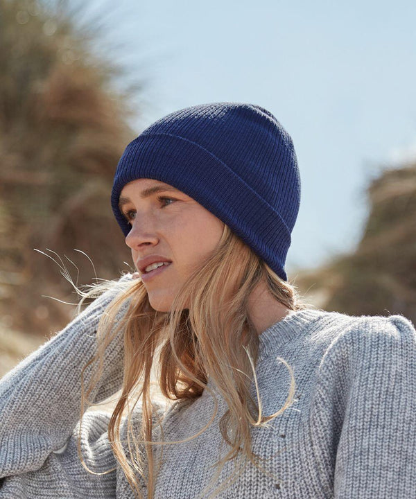 A person with long blonde hair wearing a blue Beechfield BC51N and a gray knit sweater looks into the distance with a backdrop of a natural setting featuring blurred foliage and a blue sky.