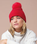 A person with shoulder-length blonde hair wears a white polo-neck jumper and the Snow Luxe Beanie by Beechfield, featuring a large red bobble, while sitting against a plain beige background.