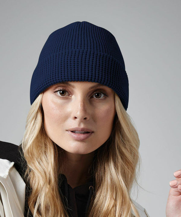 A person with long blonde hair wears a blue Beechfield BC52N knit beanie and a white jacket over a black shirt, looking directly at the camera. The background is plain grey.