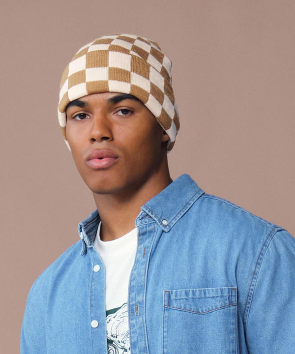 A person wears a Beechfield Checkerboard beanie in tan and white, a graphic white T-shirt, and a light blue denim button-up shirt against a plain beige background.
