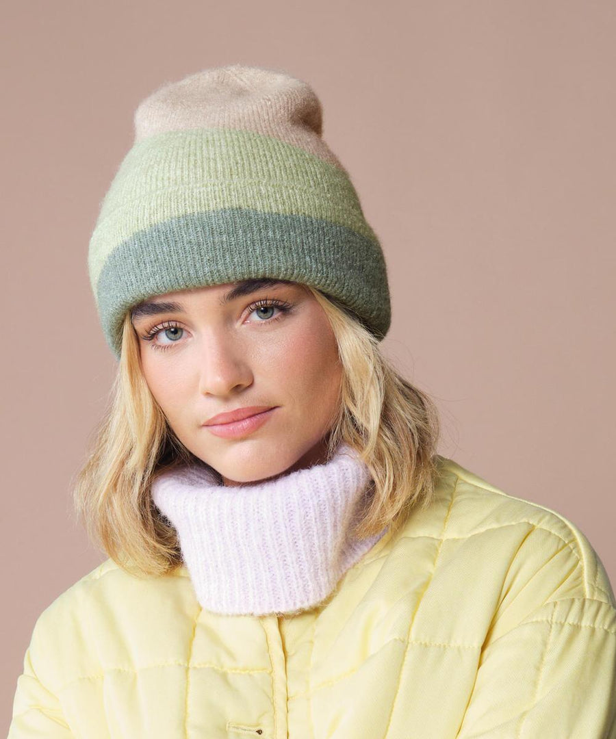 A person with shoulder-length blonde hair wears the Beechfield Soft Wave Beanie in green and beige, paired with a pale yellow puffer jacket and a thick light purple polo-neck scarf, set against a plain light beige background.