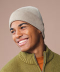 A person smiles and looks to the side against a plain beige background, wearing a Beechfield dip-dye beanie and a green zip-up jumper over an orange shirt.