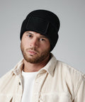 A person with a beard wearing a black Beechfield BC540 beanie and a light-colored, textured button-up shirt over a white top is staring directly at the camera with a neutral expression. The background is plain gray.