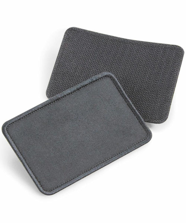 Two rectangles of black Beechfield BC600 elbow brace pads are displayed. One pad is positioned on top of the other, slightly offset, showcasing different textures. Both pads have rounded corners and visible seams around the edges.