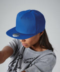 A person wearing a royal blue Beechfield BC610 flat-brimmed baseball cap and a gray T-shirt with black line art on it. The person is looking down, partially obscuring their face. The background is plain and light gray.