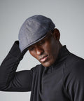 A person wearing a gray Beechfield BC621 flat cap and a black zip-up sweater is looking down, with one hand touching the cap. The background is plain and gray.