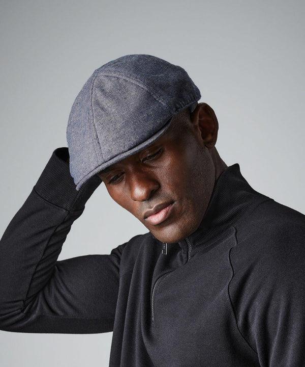 A person wearing a gray Beechfield BC621 flat cap and a black zip-up sweater is looking down, with one hand touching the cap. The background is plain and gray.