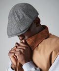 A person wearing a gray flat cap and a beige jacket with a brown corduroy collar is looking downward. The individual is holding part of the Beechfield BC622 up to their face, covering the mouth area. The background is plain gray.