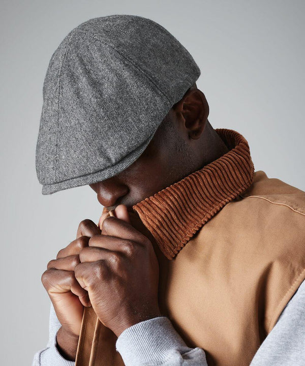 A person wearing a gray flat cap and a beige jacket with a brown corduroy collar is looking downward. The individual is holding part of the Beechfield BC622 up to their face, covering the mouth area. The background is plain gray.