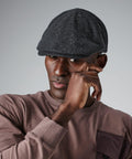 A person wearing a gray Beechfield BC623 flat cap and a mauve sweatshirt with a front pocket touches their face. The background is plain gray.