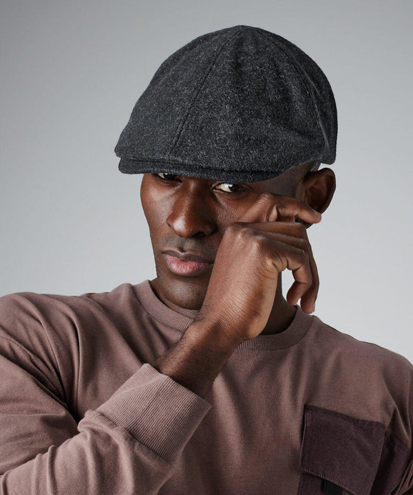 A person wearing a gray Beechfield BC623 flat cap and a mauve sweatshirt with a front pocket touches their face. The background is plain gray.