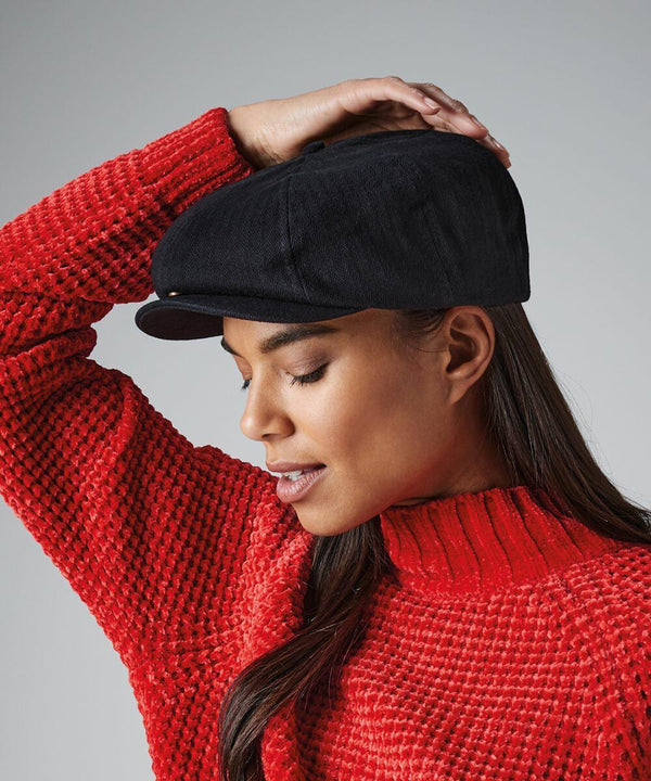 A side view of a person wearing a black Beechfield BC624 flat cap and a chunky red sweater. They have long brown hair and are holding the brim of the cap with their right hand. The background is plain gray, highlighting the person and their attire.
