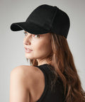 A woman with long, wavy brown hair is looking over her shoulder at the camera. She is wearing a black Beechfield BC62N and a black tank top, set against a plain, light gray background.