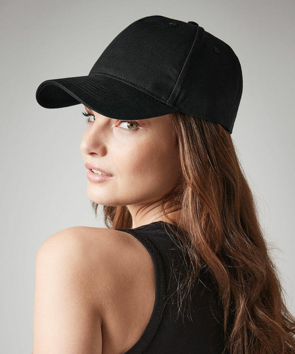 A woman with long, wavy brown hair is looking over her shoulder at the camera. She is wearing a black Beechfield BC62N and a black tank top, set against a plain, light gray background.