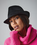 A woman wearing a black fedora hat and a bright pink knitted sweater poses against a grey background. She has chin-length hair and holds one hand to her face. Her expression is calm and thoughtful as she wears the BC630 by Beechfield.