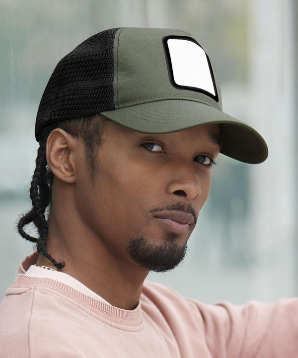 A person with a short beard and plaits wears a green and black Beechfield appliqué patch 5-panel trucker hat featuring a blank white patch on the front, paired with a light pink crew-neck shirt, looking at the camera neutrally.