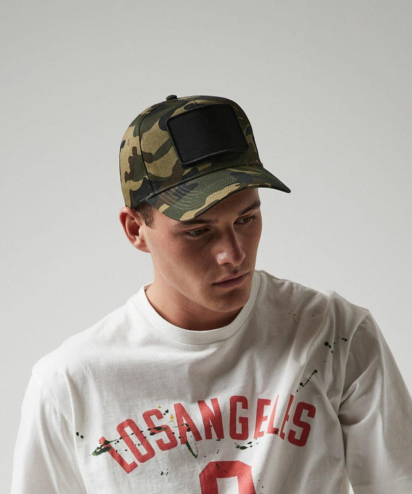 A young man is wearing a camouflage Beechfield BC638 with a black patch on the front and a white t-shirt with "LOS ANGELES" and a red number "8" printed on it. He is looking down with a neutral expression against a plain background.