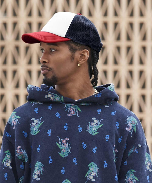 A man in a Beechfield Retro 5-panel trucker cap and a blue floral hoodie stands before a beige geometric wall, looking left. He has plaited hair and gold hoop earrings.
