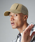A person with short hair is wearing a beige Beechfield BC641 baseball cap with a mesh back and an ear piercing. They are dressed in a gray plaid shirt and a white t-shirt underneath. The person looks to the side with a slightly surprised expression, raising one hand near their face.