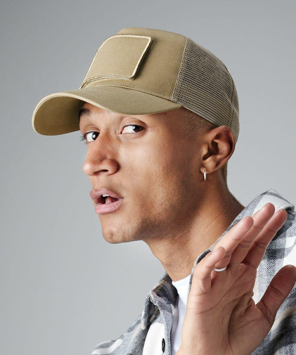 A person with short hair is wearing a beige Beechfield BC641 baseball cap with a mesh back and an ear piercing. They are dressed in a gray plaid shirt and a white t-shirt underneath. The person looks to the side with a slightly surprised expression, raising one hand near their face.