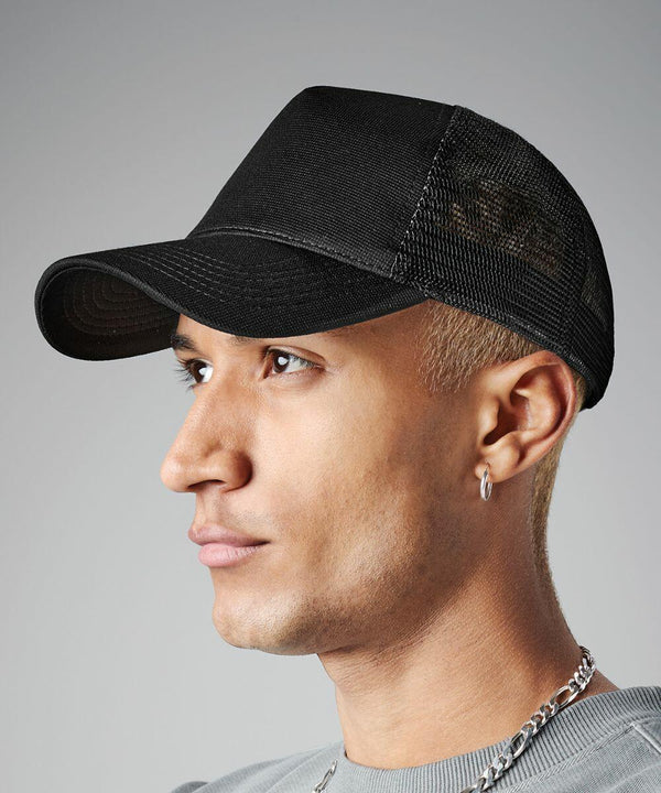 A close-up side profile of a person wearing a black Beechfield BC642 baseball cap with a mesh back. They have short, blond hair and are wearing a silver hoop earring and a silver chain necklace. The background is plain gray.