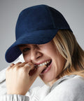 A person wearing a navy blue Beechfield BC643 cap and a light gray top smiles and playfully bites a finger. The individual's blonde hair is partially visible under the cap, and they are wearing two rings on their fingers. The background is a neutral gray.
